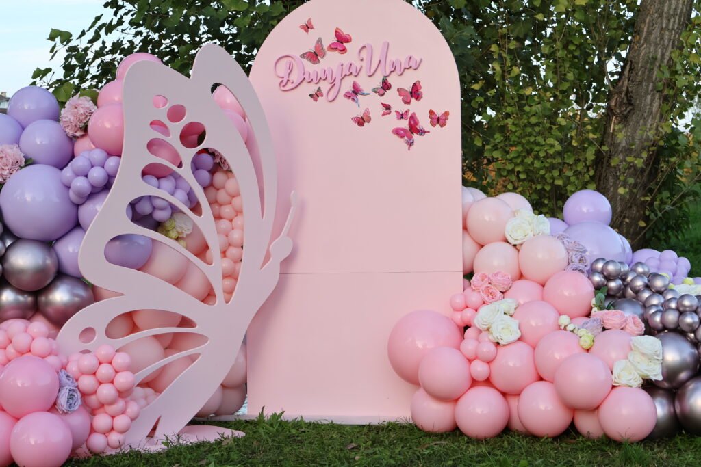 Raskošna roza drvena pozadina u obliku luka s velikim 3D leptirom i personaliziranim imenom, okružena bogatim organskim girlandama od pastelnih rozih, lila i srebrnih balona te svježim cvijećem za rođendansku proslavu na otvorenom, Joli Spot Zagreb